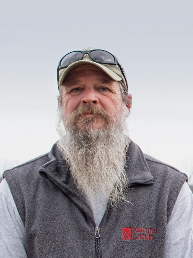 a man with a mustache and long gray beard with a baseball hat with sunglasses perched on top and wearing a gray shirt-less fleece over a long-sleeve gray shirt