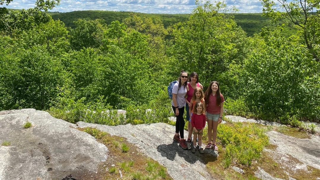A family stands on a large gray boulder in front of a blue sky with white clouds and green trees