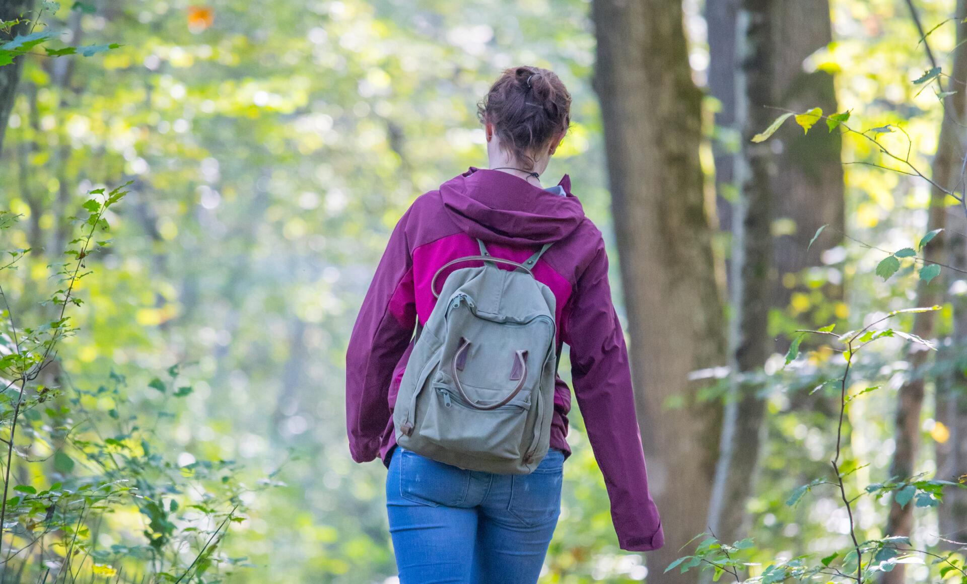 girl with purple jacket and gray backpack walking through the woods