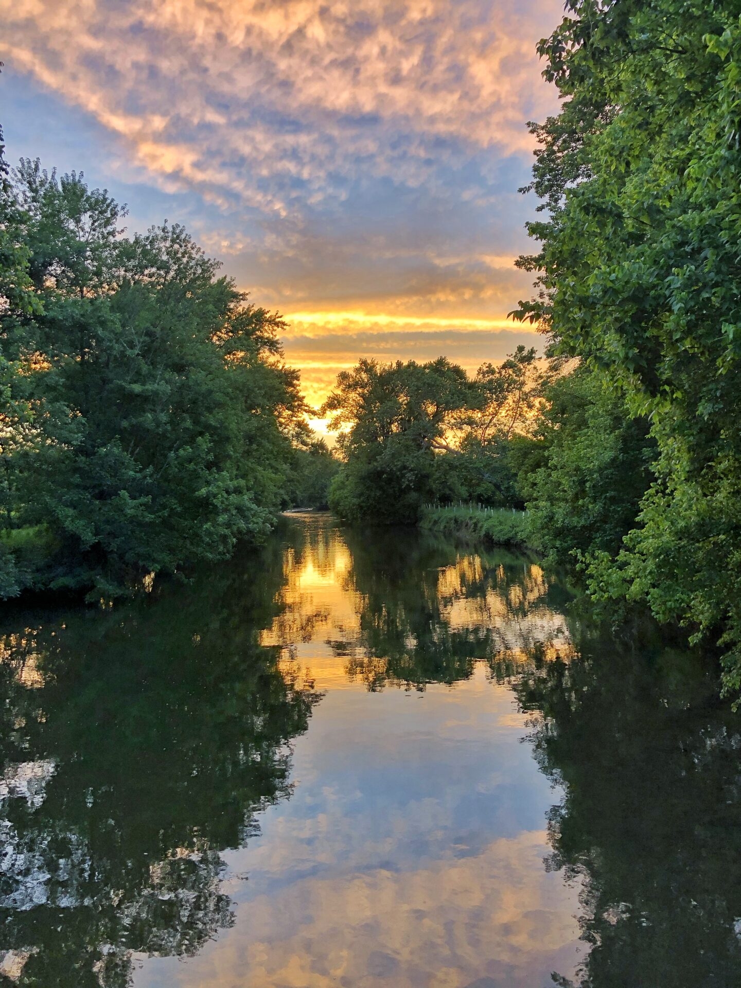 a sunset in the sky and reflected on the water of a creek that has trees on both sides