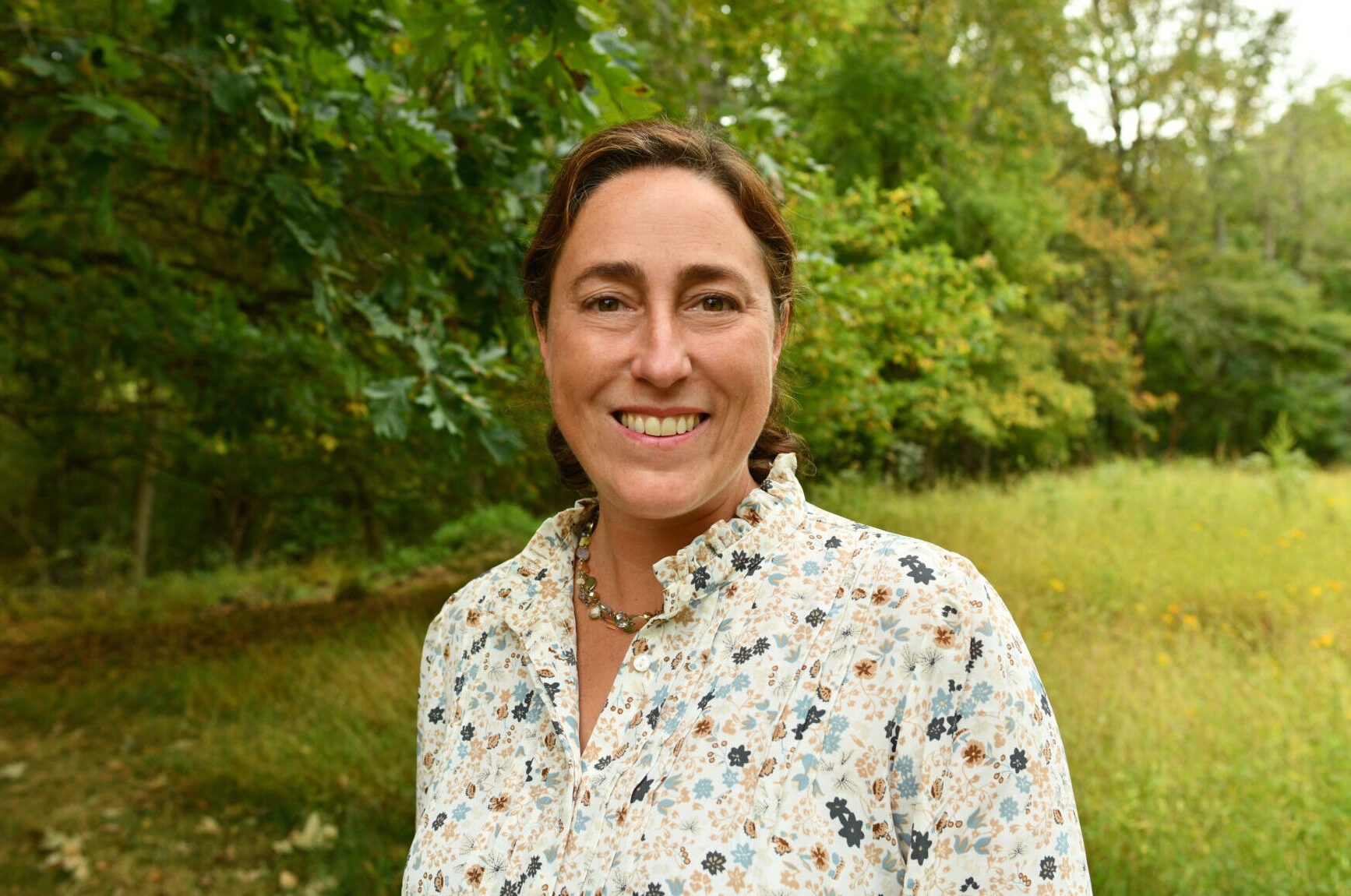 a smiling woman wearing a necklace with a floral-print shirt standing in a field with trees