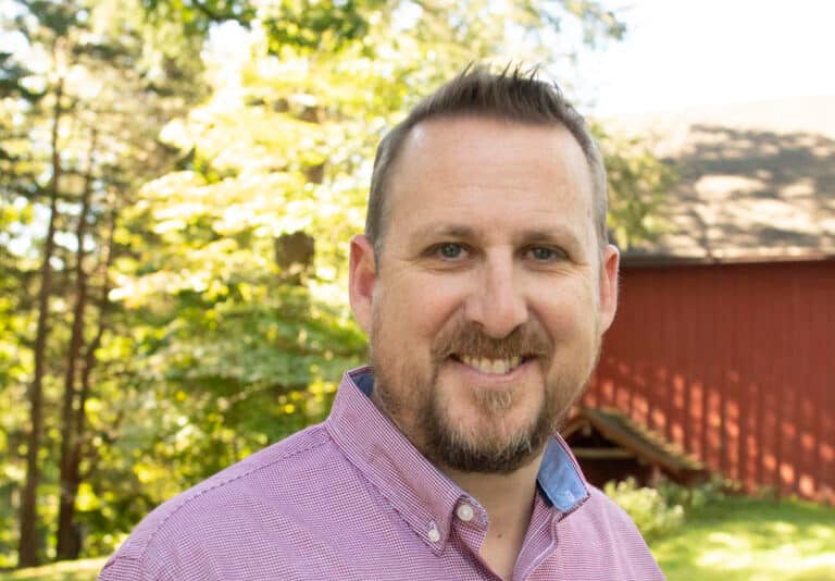 a man with a mustache and beard smiling at the camera with a red barn and trees in the background