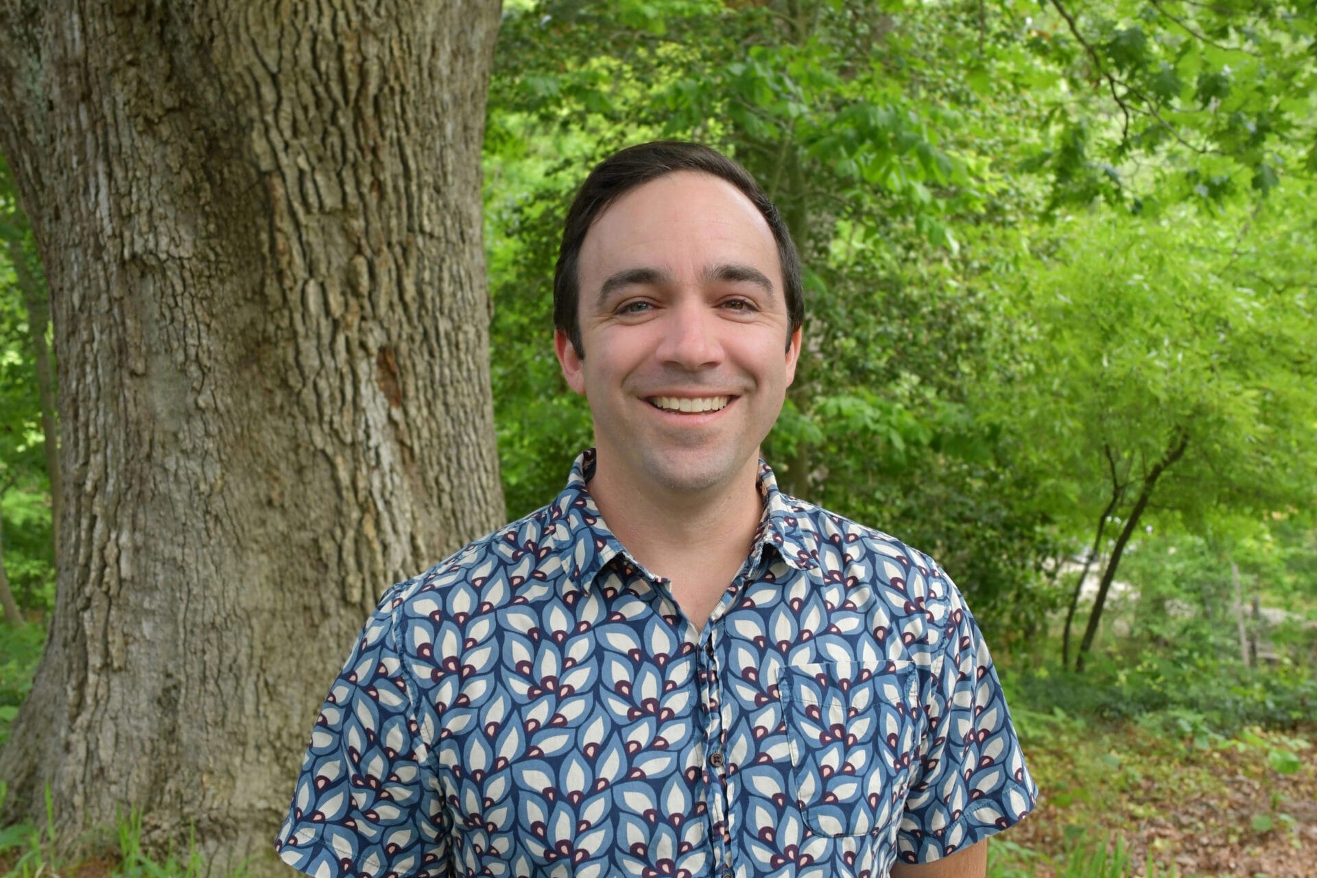 a smiling man with a blue and white patterned shirt on with trees in the background